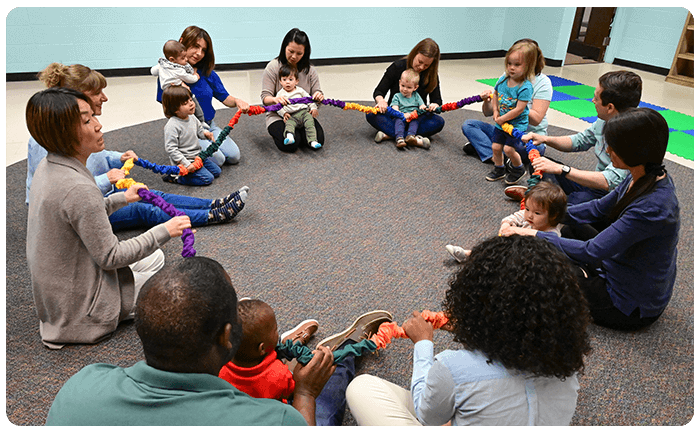Parents and children in Music Together class sitting on rug in circle holding a stretchy band